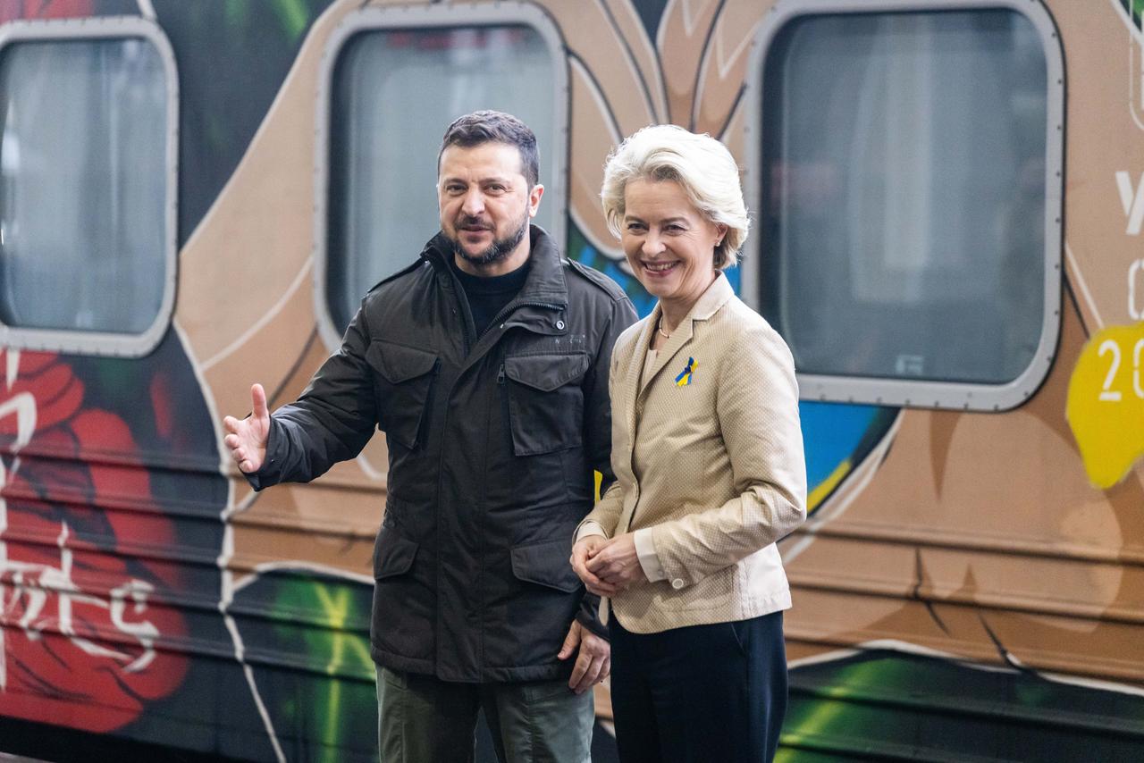 Selenskyj und von der Leyen stehen nebeneinander und schauen in die Kamera. Selenskyj gestikuliert mit einer Hand. Im Hintergrund ist ein Zug zu sehen.  
