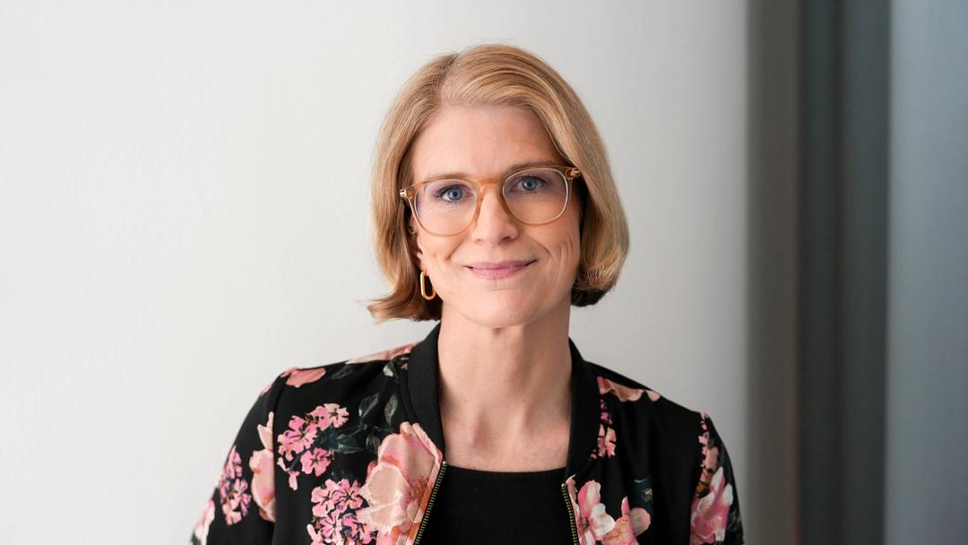 Portrait einer Frau mit blonden, kinnlangen Haaren und Brille, die lächelnd mit geneigtem Kopf in die Kamera blickt.