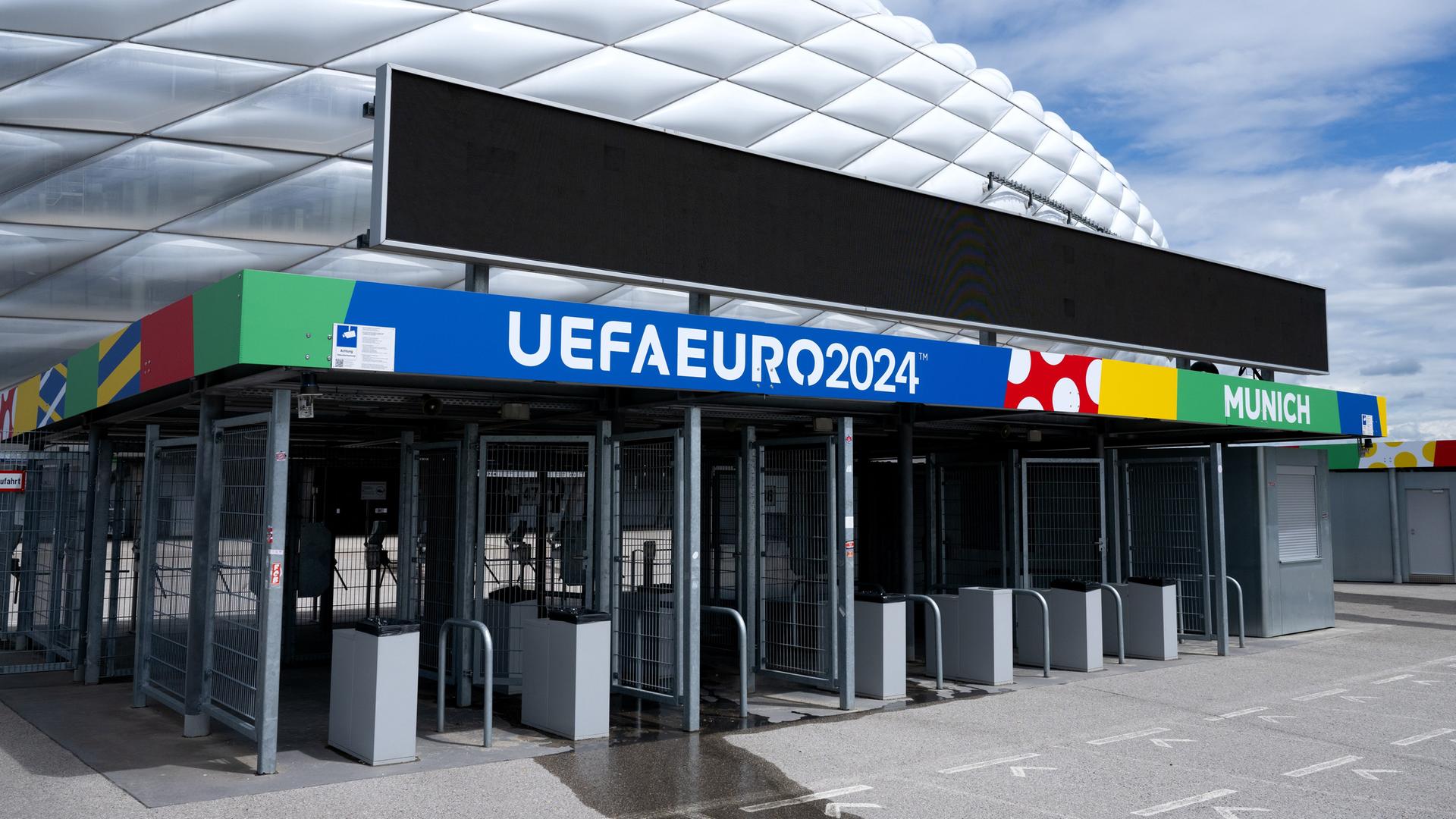 Zu sehen ist das Fußball-Stadion in München. Im Vordergrund ein buntes Schild mit der Aufschrift "UEFA EURO 2024". Zu sehen ist das Fußball-Stadion in München. Im Vordergrund ein buntes Schild mit der Aufschrift "UEFA EURO 2024".