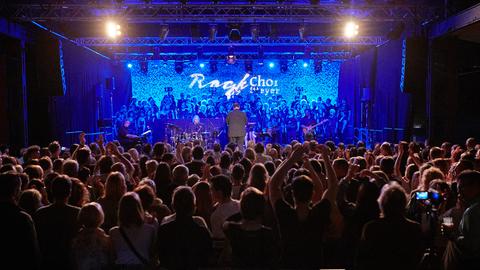 Auf einer blau eingeleuchteten Bühne stehen der Chor und die Band. Vor der Bühne steht dichtgedrängt das Publikum.