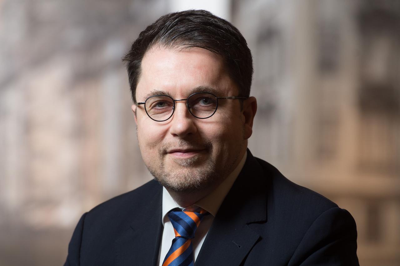 Ein Mann in Anzug und Krawatte, mit Seitenscheitel, Brille und Drei-Tage-Bart blickt in die Kamera.