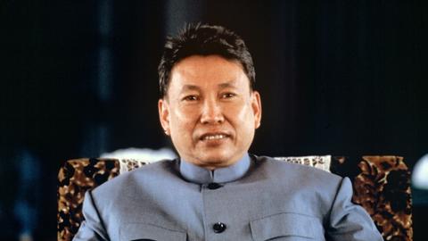 Pol Pot sitzt mit einer blauen Jacke auf einem Stuhl. Er hat eine kurze Frisur und blickt in die Kamera.