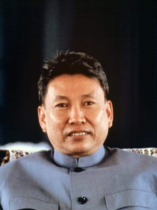 Pol Pot sitzt mit einer blauen Jacke auf einem Stuhl. Er hat eine kurze Frisur und blickt in die Kamera.