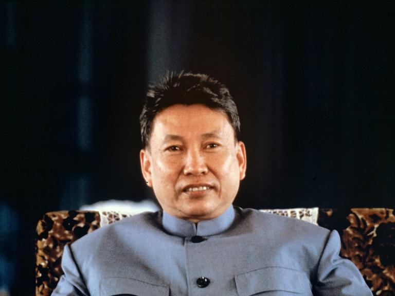 Pol Pot sitzt mit einer blauen Jacke auf einem Stuhl. Er hat eine kurze Frisur und blickt in die Kamera.