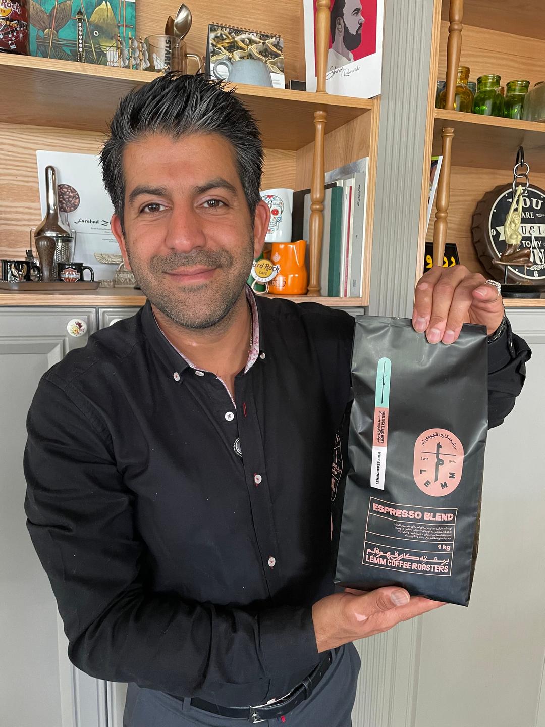 Vor einem Regal steht ein Mann mit schwarzen Haaren und schwarzem Hemd und hebt eine Kaffeepackung hoch.