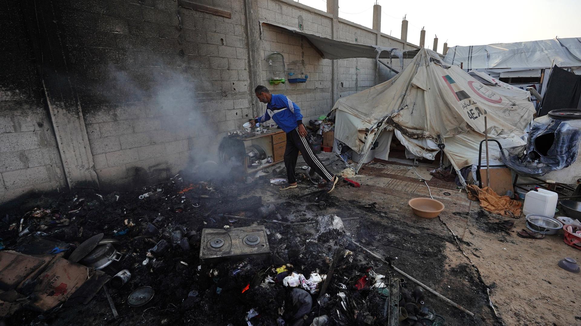 Aus der Asche eines zerstörten Zeltes neben einer Mauer steigt Rauch auf, ein Mann sichert noch brauchbare Gegenstände. Im Hintergrund steht ein weitgehend intaktes Zelt. 