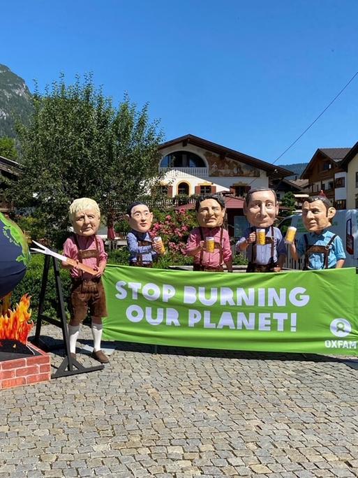 Aktivisten von Oxfam protestieren mit Politiker-Köpfen am Rande des G7-Gipfels auf Schloss Elmau gegen die Klimapolitik der Industrienationen. Die Figuren Scholz. Biden und Johnson drehen dabei eine Weltkugel aus Pappmaché über einer Feuerstelle aus Pappe.