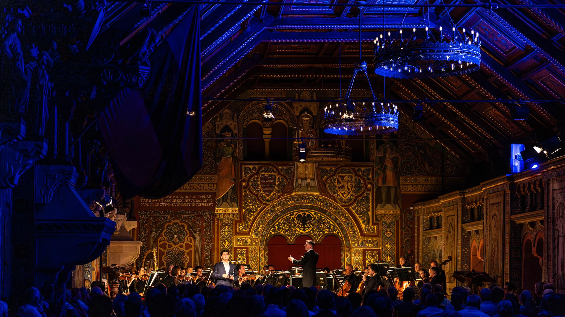 Orchesterkonzert in prachtvollem historischen Saal: Dirigent und Solist vor der Meininger Hofkapelle, umgeben von reich verzierter Architektur, warmem Goldlicht und großen Kronleuchtern, das Publikum dicht gefüllt im Vordergrund. Orchesterkonzert in prachtvollem historischen Saal: Dirigent und Solist vor der Meininger Hofkapelle, umgeben von reich verzierter Architektur, warmem Goldlicht und großen Kronleuchtern, das Publikum dicht gefüllt im Vordergrund.