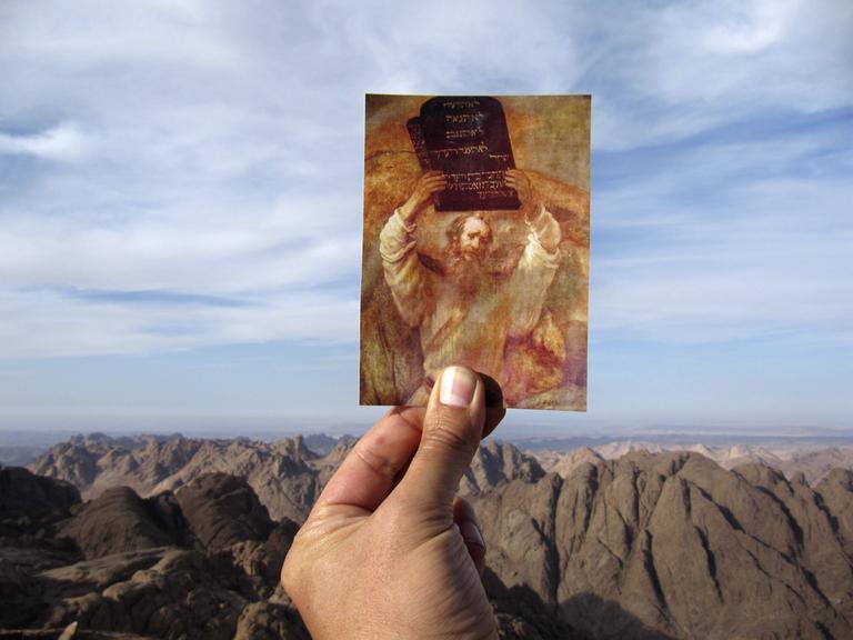 Eine Hand hält vor einem Gebirge eine Postkarte hoch, auf der ein Rubens Gemälde zu sehen ist, das Moses mit der Tafel der zehn Gebote zeigt.