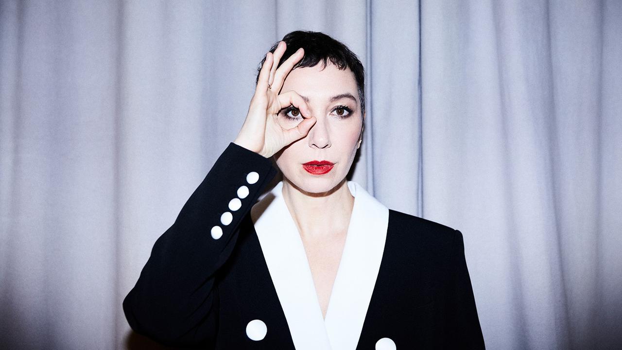 Eine Frau mit weiß geschminktem Gesicht und roten Lippen trägt ein schwarzes Jackett mit weißen Knöpfen und weißem Kragen. Sie steht vor einem hellen Vorhang und blickt mit ihrem rechten Auge durch einen Ring, den sie mit Daumen und Zeigefinger formt.