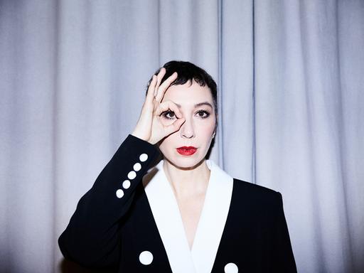 Eine Frau mit weiß geschminktem Gesicht und roten Lippen trägt ein schwarzes Jackett mit weißen Knöpfen und weißem Kragen. Sie steht vor einem hellen Vorhang und blickt mit ihrem rechten Auge durch einen Ring, den sie mit Daumen und Zeigefinger formt.