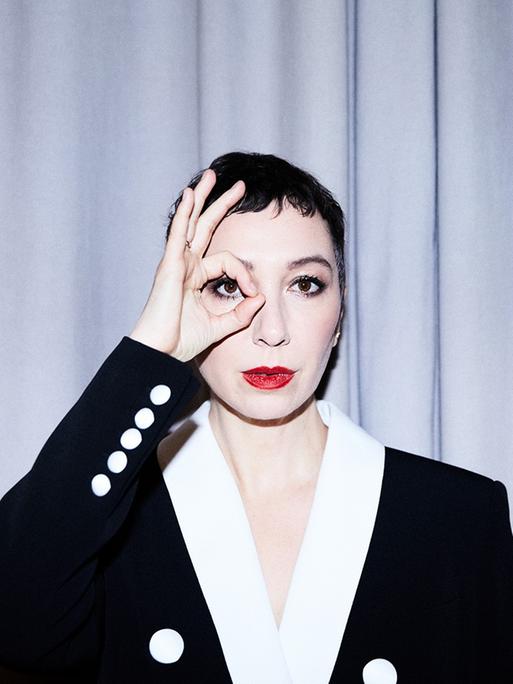 Eine Frau mit weiß geschminktem Gesicht und roten Lippen trägt ein schwarzes Jackett mit weißen Knöpfen und weißem Kragen. Sie steht vor einem hellen Vorhang und blickt mit ihrem rechten Auge durch einen Ring, den sie mit Daumen und Zeigefinger formt.