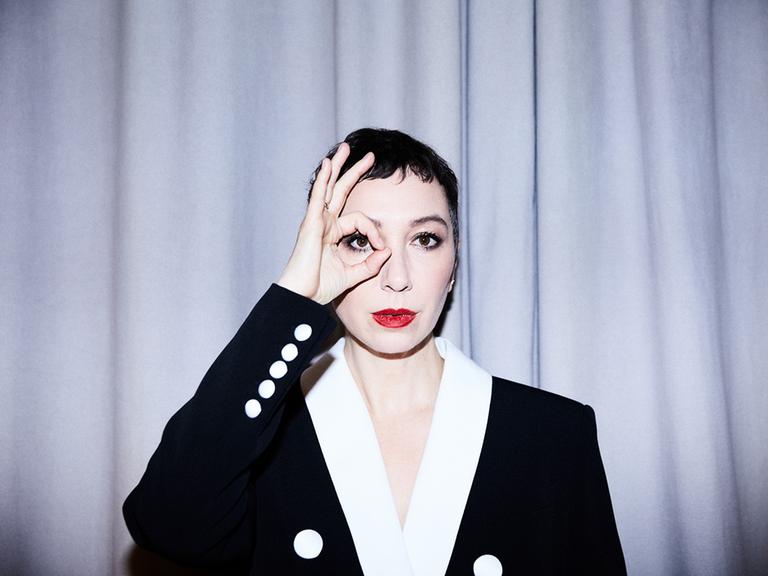 Eine Frau mit weiß geschminktem Gesicht und roten Lippen trägt ein schwarzes Jackett mit weißen Knöpfen und weißem Kragen. Sie steht vor einem hellen Vorhang und blickt mit ihrem rechten Auge durch einen Ring, den sie mit Daumen und Zeigefinger formt.