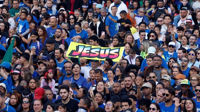 In einem Stadion sitzen unzählige Menschen, in der Mitte wird ein Banner mit der Aufschrift "Jesus" in die Luft.