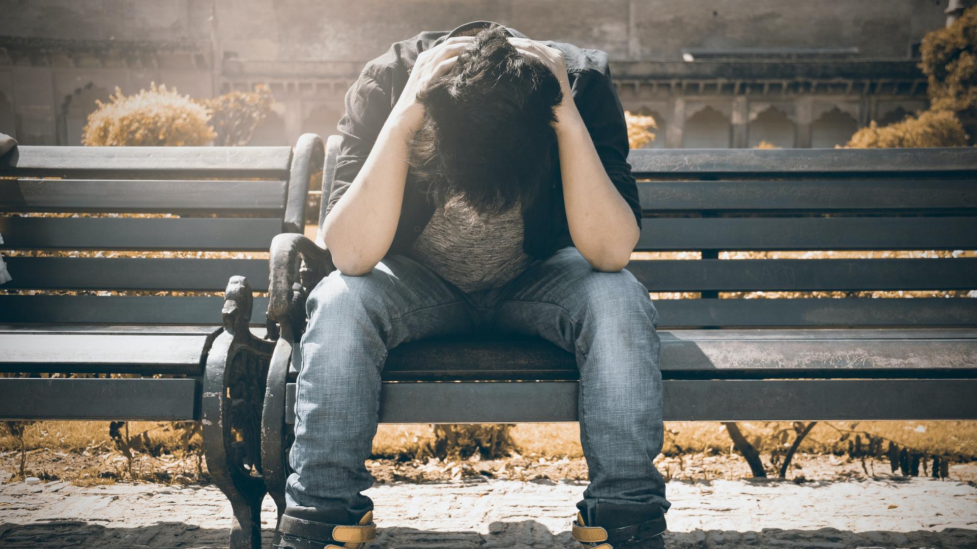 Mann hält Kopf in den Händen und sitzt kaputt auf einer Bank