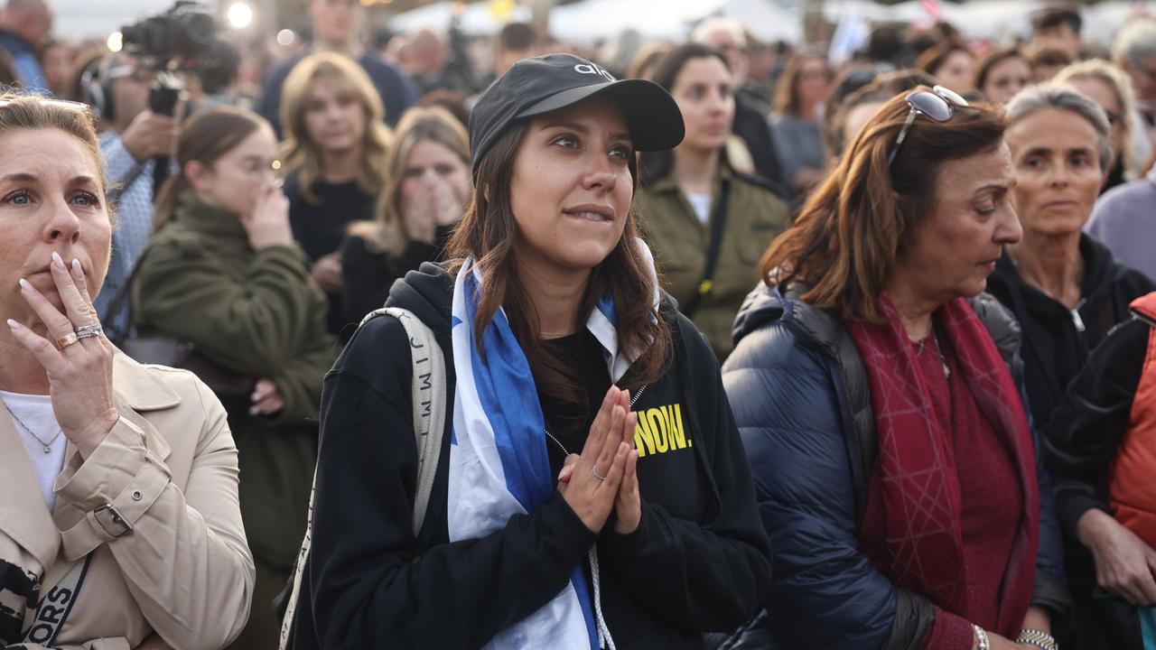 Eine Gruppe von Menschen steht auf einem Platz. Im Zentrum des Bildes ist eine Frau zu sehen, die eine israelische Flagge um die Schultern trägt und angespannt und hoffnungsvoll, die Handflächen aufeinander gelegt, nach vorn schaut.