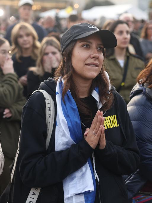 Eine Gruppe von Menschen steht auf einem Platz. Im Zentrum des Bildes ist eine Frau zu sehen, die eine israelische Flagge um die Schultern trägt und angespannt und hoffnungsvoll, die Handflächen aufeinander gelegt, nach vorn schaut.