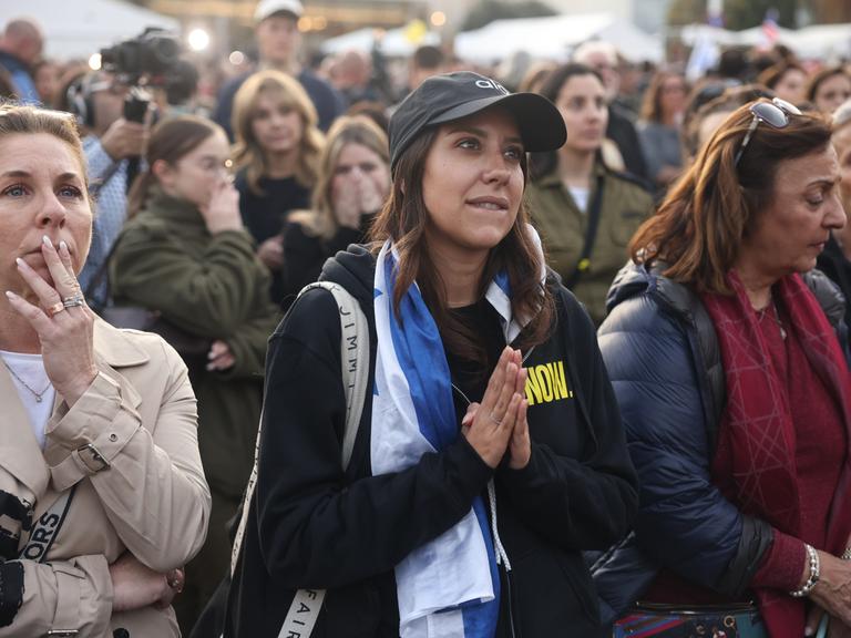 Eine Gruppe von Menschen steht auf einem Platz. Im Zentrum des Bildes ist eine Frau zu sehen, die eine israelische Flagge um die Schultern trägt und angespannt und hoffnungsvoll, die Handflächen aufeinander gelegt, nach vorn schaut.