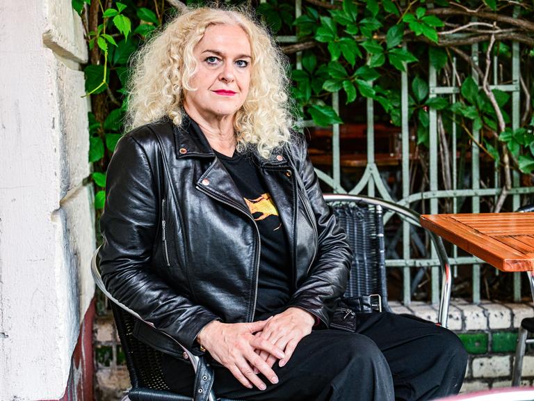 Vernesa Berbo posiert auf einem Stuhl sitzend für die Kamera. Sie trägt eine schwarze Lederjacke, roten Lippenstift und wilde blonde Locken.
