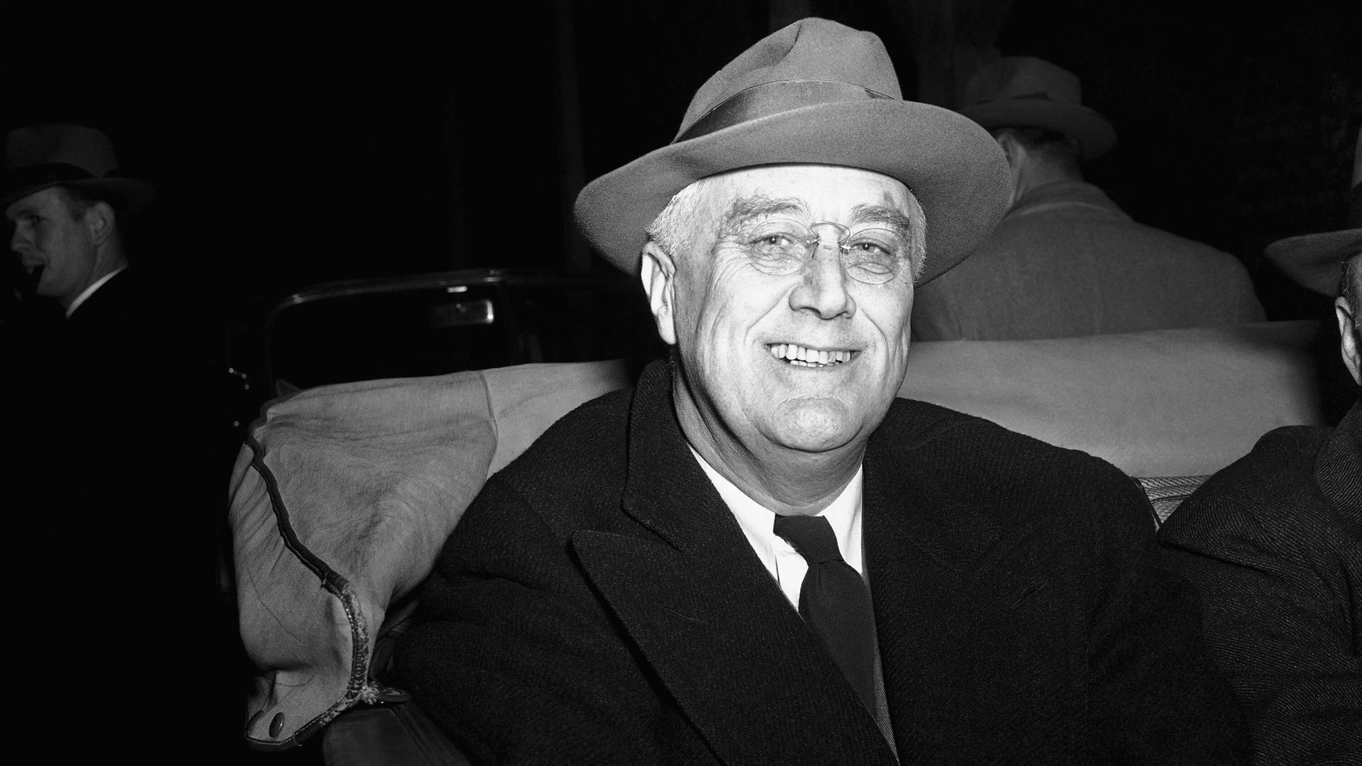 Franklin D. Roosevelt lächelt auf einem Schwarz-Weiß-Foto mit Hut nach eine Feier mit Nachbarn in die Kamera. Franklin D. Roosevelt lächelt auf einem Schwarz-Weiß-Foto mit Hut nach eine Feier mit Nachbarn in die Kamera.