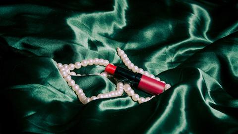 Ein Lippenstift und eine Perlenkette liegen auf einem grünen Tuch.