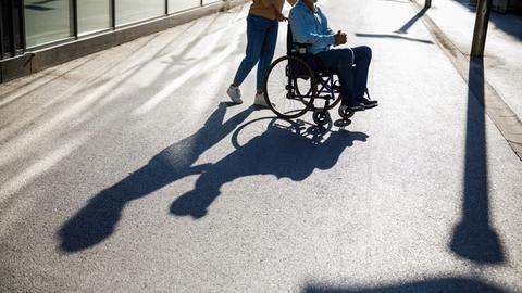 Eine Person schiebt eine andere Person in einem Rollstuhl. Ihr Schatten ist auf der Strasse sichtbar.