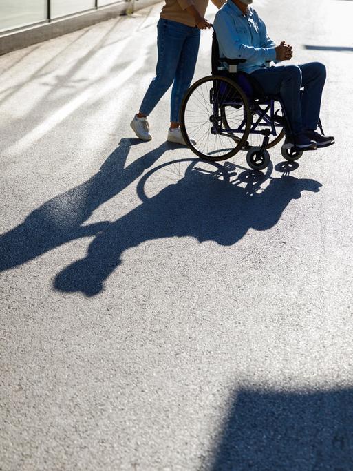 Eine Person schiebt eine andere Person in einem Rollstuhl. Ihr Schatten ist auf der Strasse sichtbar.