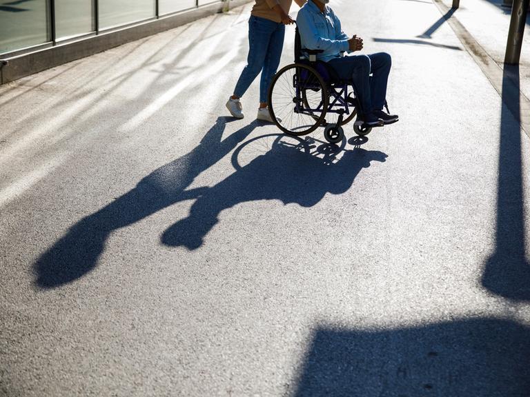 Eine Person schiebt eine andere Person in einem Rollstuhl. Ihr Schatten ist auf der Strasse sichtbar.