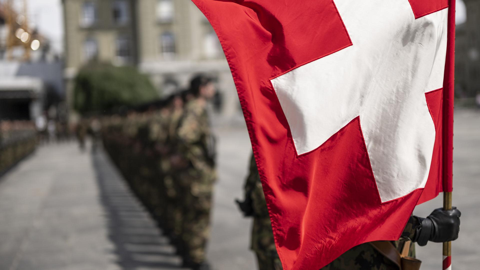 Eine Truppe der Schweizer Armee steht auf einem Marktplatz, die vordere Person hält eine Schweizer Flagge. Eine Truppe der Schweizer Armee steht auf einem Marktplatz, die vordere Person hält eine Schweizer Flagge.