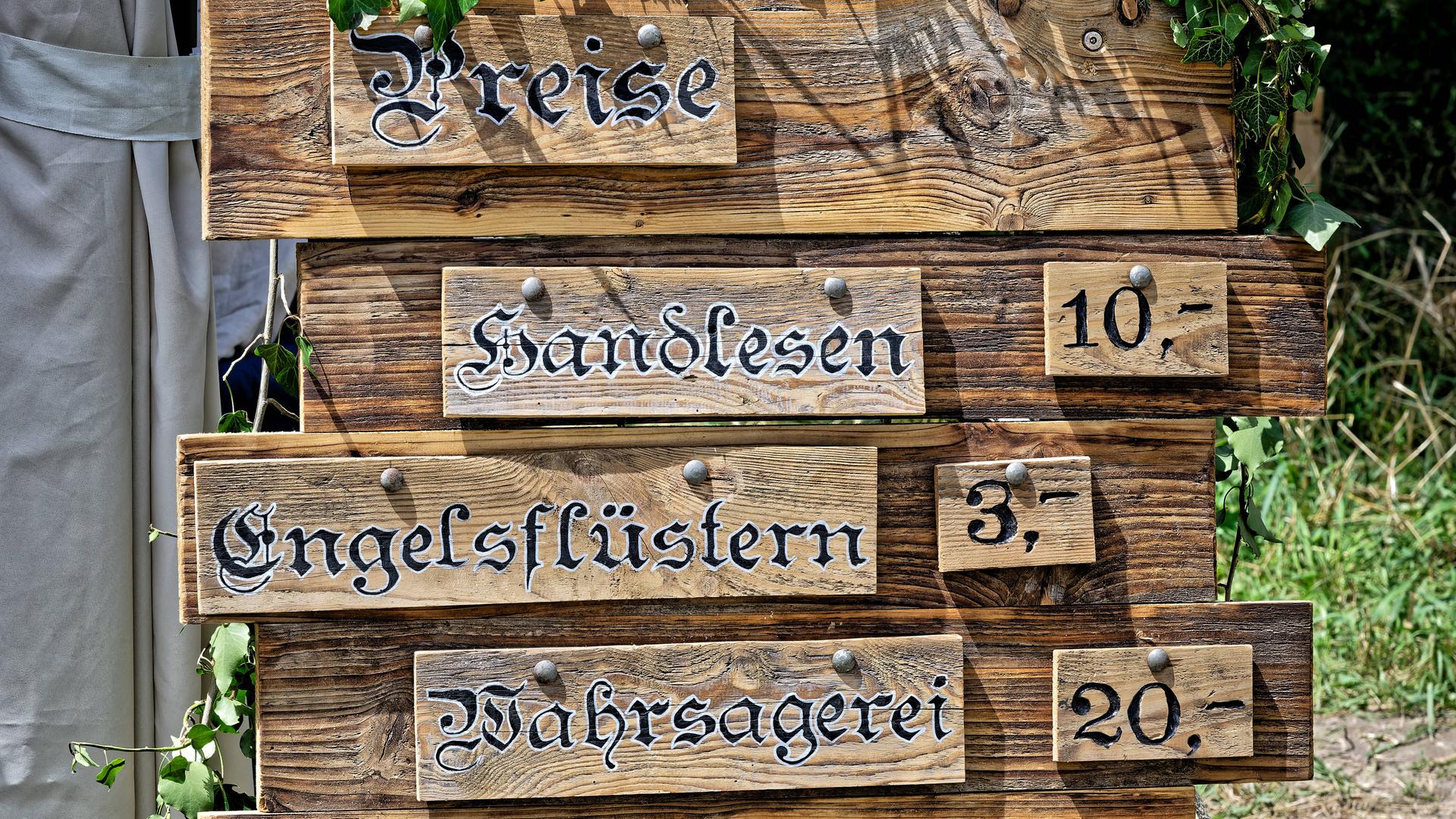 Auf dem Bild ist eine Holztafel abgebildet, auf der die Preise für folgende Leistungen aufgelistet sind: Handlesen, Engelsflüstern und Wahrsagerei Auf dem Bild ist eine Holztafel abgebildet, auf der die Preise für folgende Leistungen aufgelistet sind: Handlesen, Engelsflüstern und Wahrsagerei