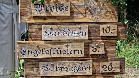 Auf dem Bild ist eine Holztafel abgebildet, auf der die Preise für folgende Leistungen aufgelistet sind: Handlesen, Engelsflüstern und Wahrsagerei Auf dem Bild ist eine Holztafel abgebildet, auf der die Preise für folgende Leistungen aufgelistet sind: Handlesen, Engelsflüstern und Wahrsagerei