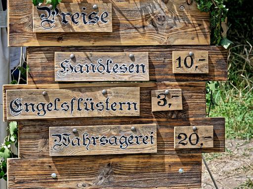 Auf dem Bild ist eine Holztafel abgebildet, auf der die Preise für folgende Leistungen aufgelistet sind: Handlesen, Engelsflüstern und Wahrsagerei