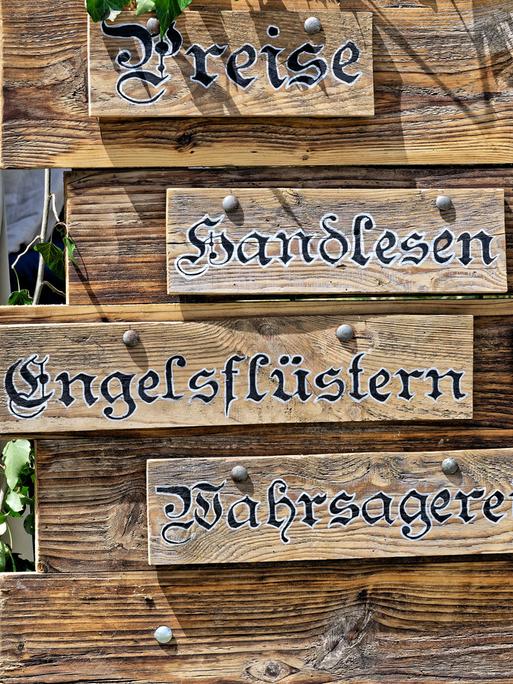 Auf dem Bild ist eine Holztafel abgebildet, auf der die Preise für folgende Leistungen aufgelistet sind: Handlesen, Engelsflüstern und Wahrsagerei