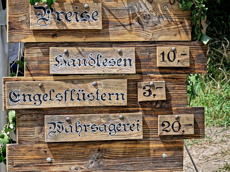 Auf dem Bild ist eine Holztafel abgebildet, auf der die Preise für folgende Leistungen aufgelistet sind: Handlesen, Engelsflüstern und Wahrsagerei