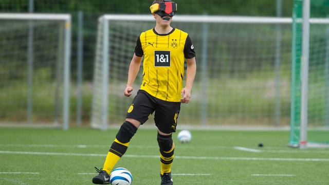 Ein Mann im Trikot von Borussia Dortmund. Er hat einen Fuß am Ball und eine Binde über den Augen. 