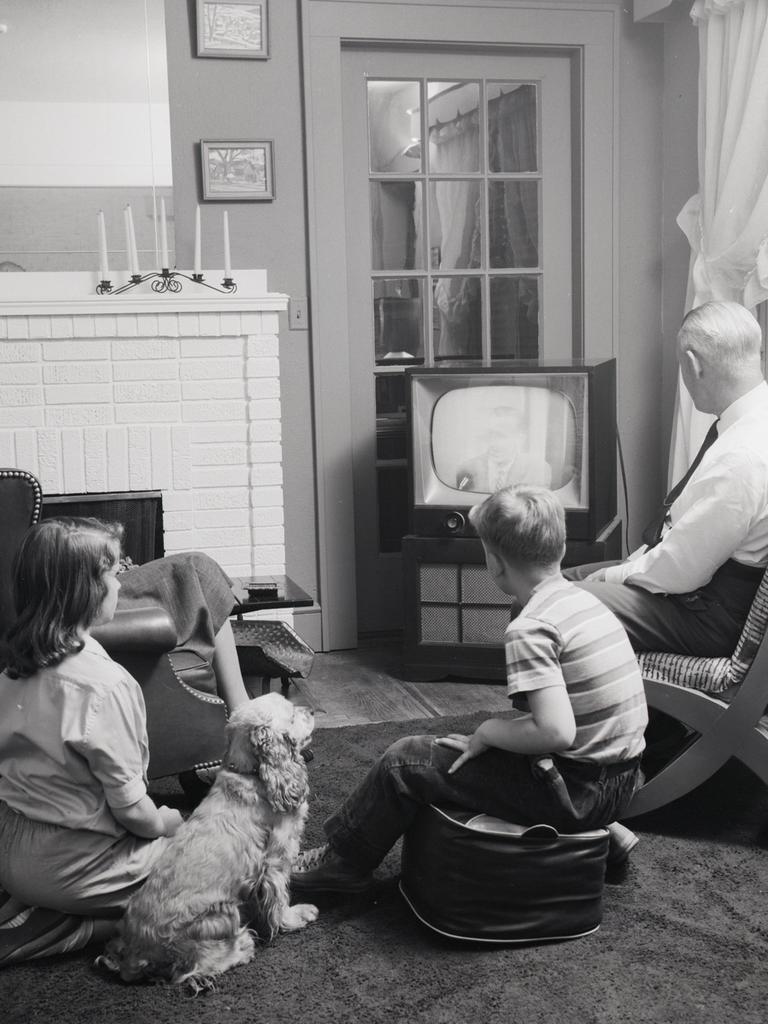 Schwarzweißfoto einer Familie mit zwei Kindern in den 1950er Jahren, die gemeinsam mit einem Hund vor dem Fernseher sitzt.