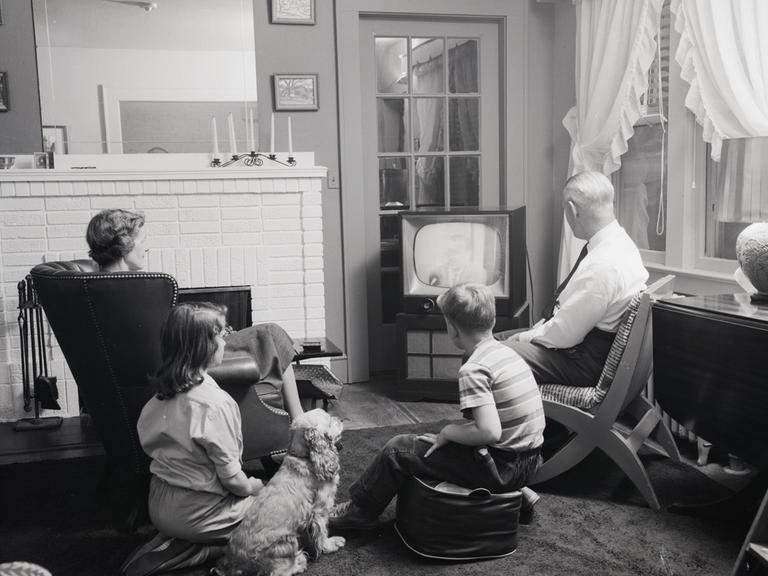 Schwarzweißfoto einer Familie mit zwei Kindern in den 1950er Jahren, die gemeinsam mit einem Hund vor dem Fernseher sitzt.