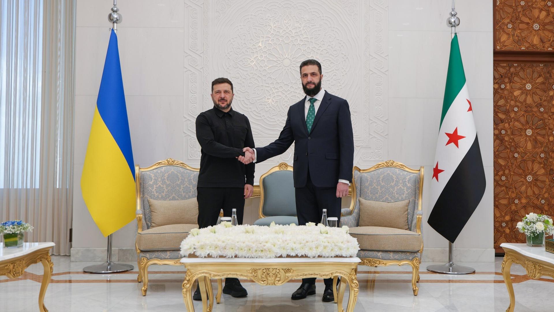 Zwei Männer in Anzügen schütteln sich die Hand. Rechts steht die ukraine Flagge, rechts die syrische.