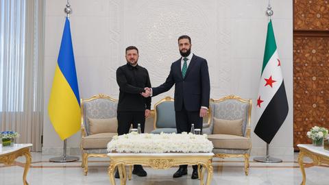 Zwei Männer in Anzügen schütteln sich die Hand. Rechts steht die ukraine Flagge, rechts die syrische.