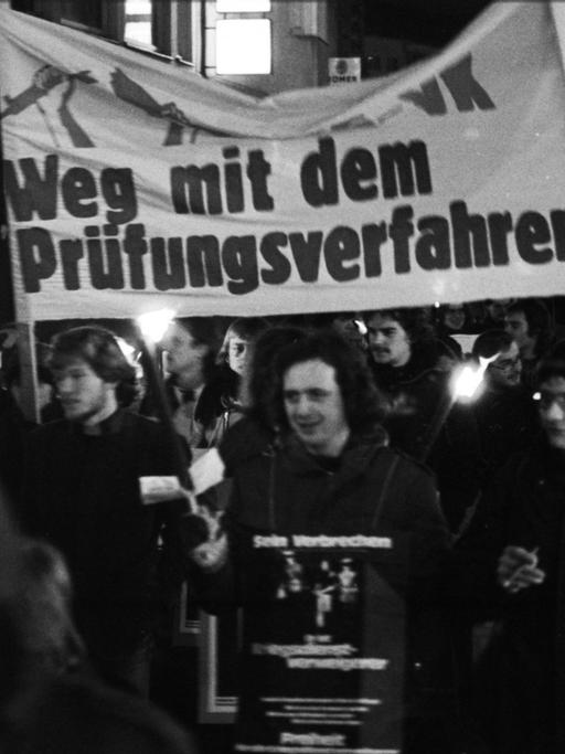 Historische schwarz-weiß-Aufnahme von einem Fackelmarsch gegen die Pruefung zur Kriegsdienstverweigerung in Bonn im Dezember 1977. Auf einem Transparent steht "Weg mit dem Prüfungsverfahren".