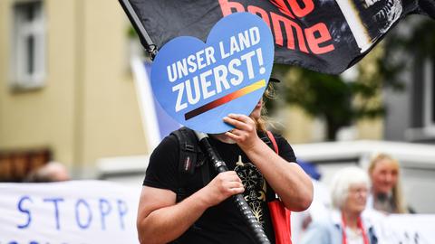 Eine Person hält auf einer Demo ein Schild in Herzform vor ihr Gesicht, auf dem auf blauem Grund steht: "Unser Land zuerst!"