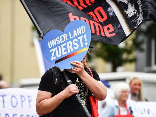 Eine Person hält auf einer Demo ein Schild in Herzform vor ihr Gesicht, auf dem auf blauem Grund steht: "Unser Land zuerst!"