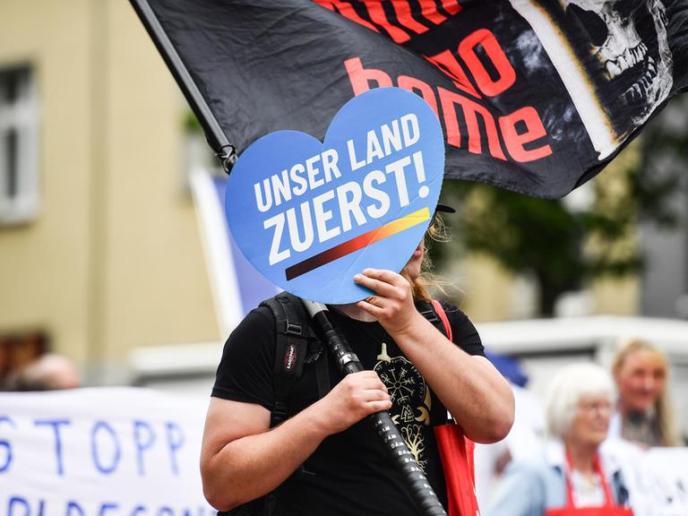 Eine Person hält auf einer Demo ein Schild in Herzform vor ihr Gesicht, auf dem auf blauem Grund steht: "Unser Land zuerst!"