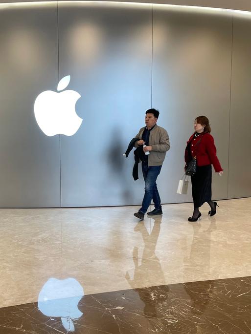 Zwei Kunden laufen durch einen Store des Apple-Konzerns. An einer Wand ist dessen Symbol in Form eines weißen Apfels mit einem Biss zu sehen. 