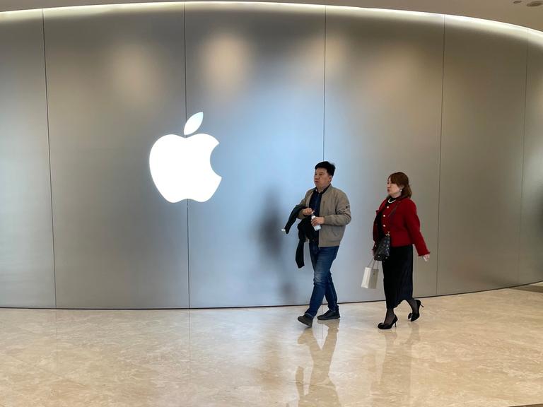 Zwei Kunden laufen durch einen Store des Apple-Konzerns. An einer Wand ist dessen Symbol in Form eines weißen Apfels mit einem Biss zu sehen. 