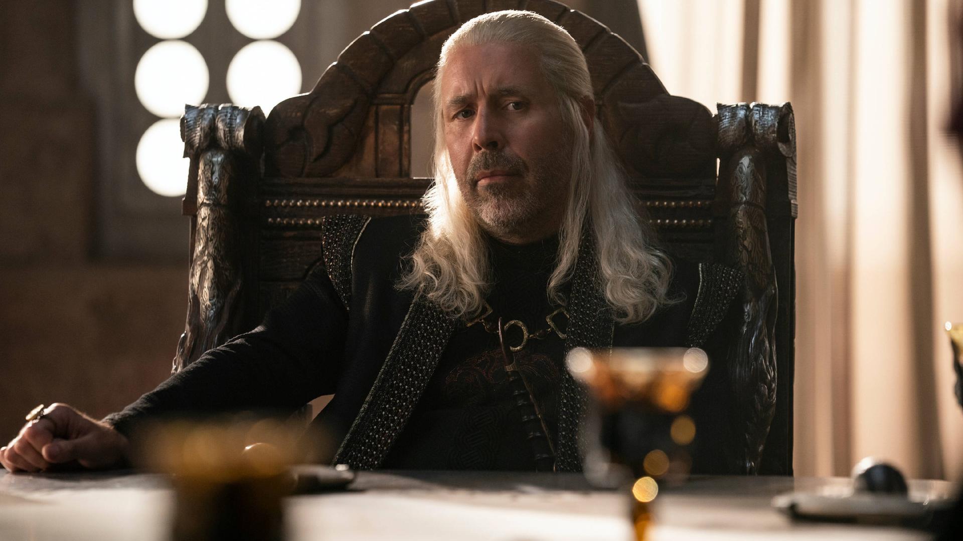 In vollem Ornat: Paddy Considine in seiner Rolle als König Viserys Targaryen in der neuen Serie "House of the Dragon". Er schaut vorsichtig, aber freundlich. Im Vordergrund sind Gläser erkennbar, er sitzt offenbar an einer Tafel. 
