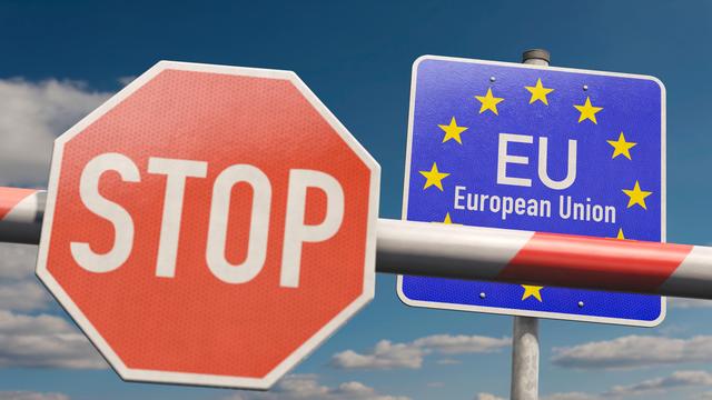 An einem Schlagbaum hängt ein Stoppschild. Dahinter steht ein EU-Schild. Beides vor einem blauen Himmel mit Wolken.