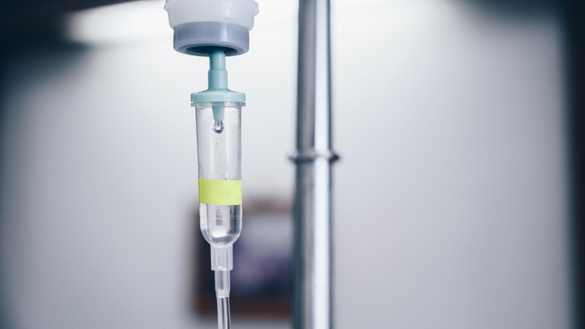 Nahaufnahme eines medizinischen Tropfs. Im Hintergrund ist ein Krankenzimmer schemenhaft zu erkennen.