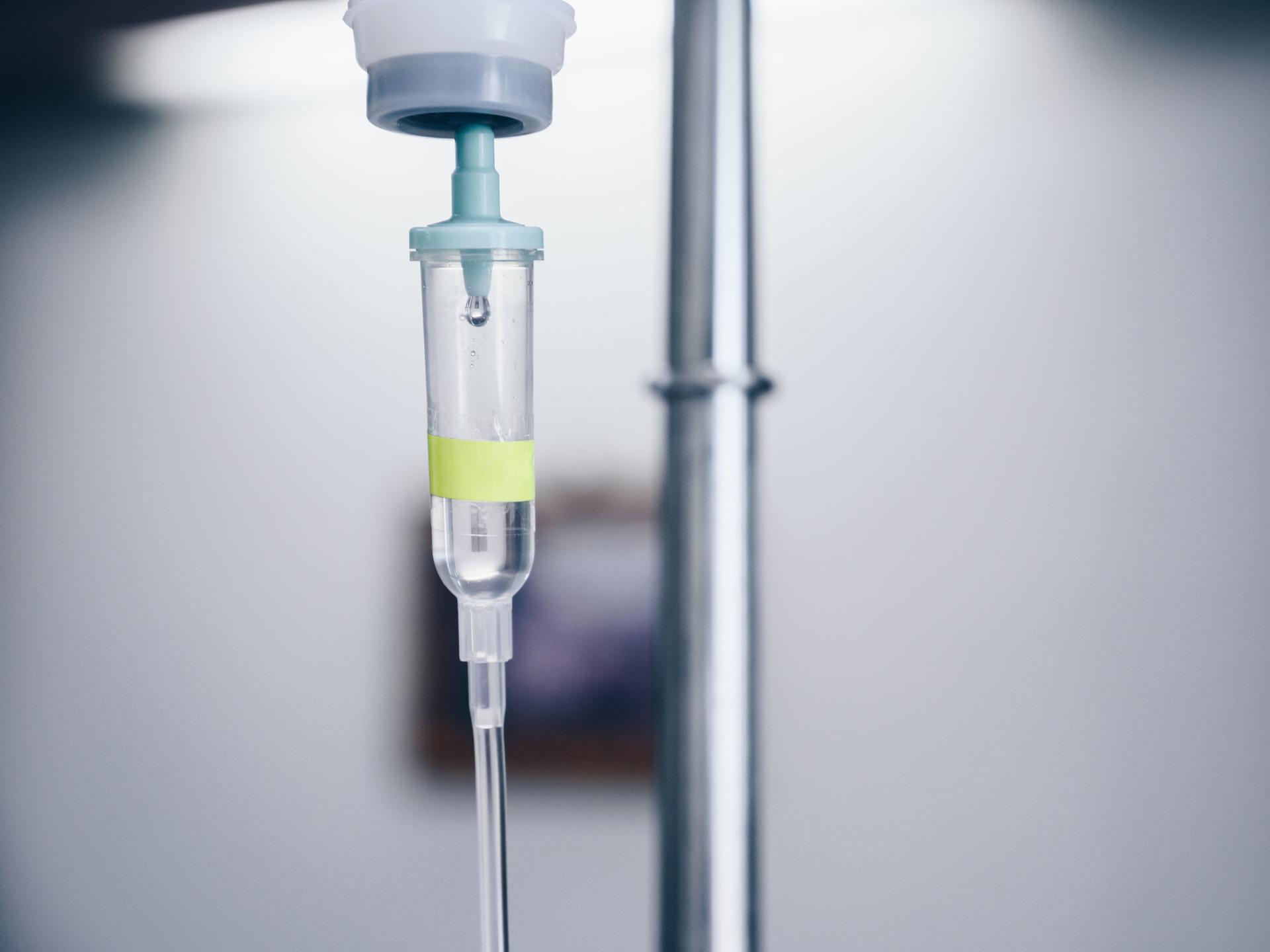 Nahaufnahme eines medizinischen Tropfs. Im Hintergrund ist ein Krankenzimmer schemenhaft zu erkennen.