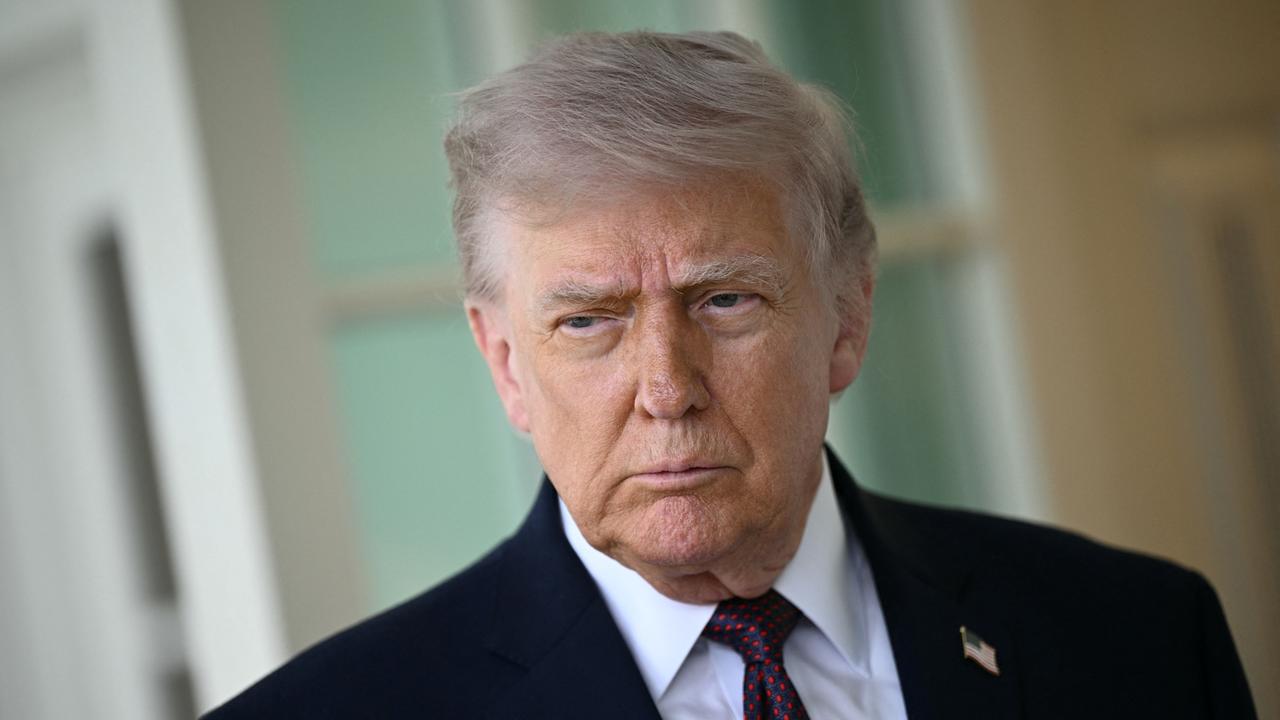 Donald Trump steht vor einer weißen Fassade mit Fenstern. Er trägt einen dunklen Anzug und Krawatte.